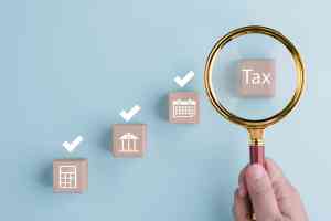 © Bussarin / stock.adobe.com Lupe vergrößert Holzklotz mit Aufschrift Tax